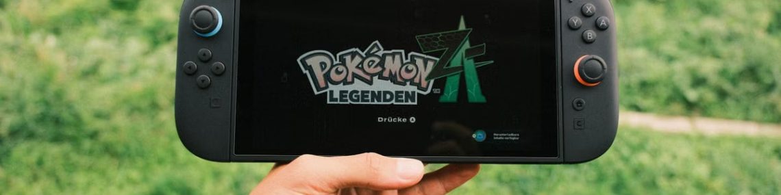 A hand holding a handheld gaming console displaying the Pokémon Legends game screen outdoors.